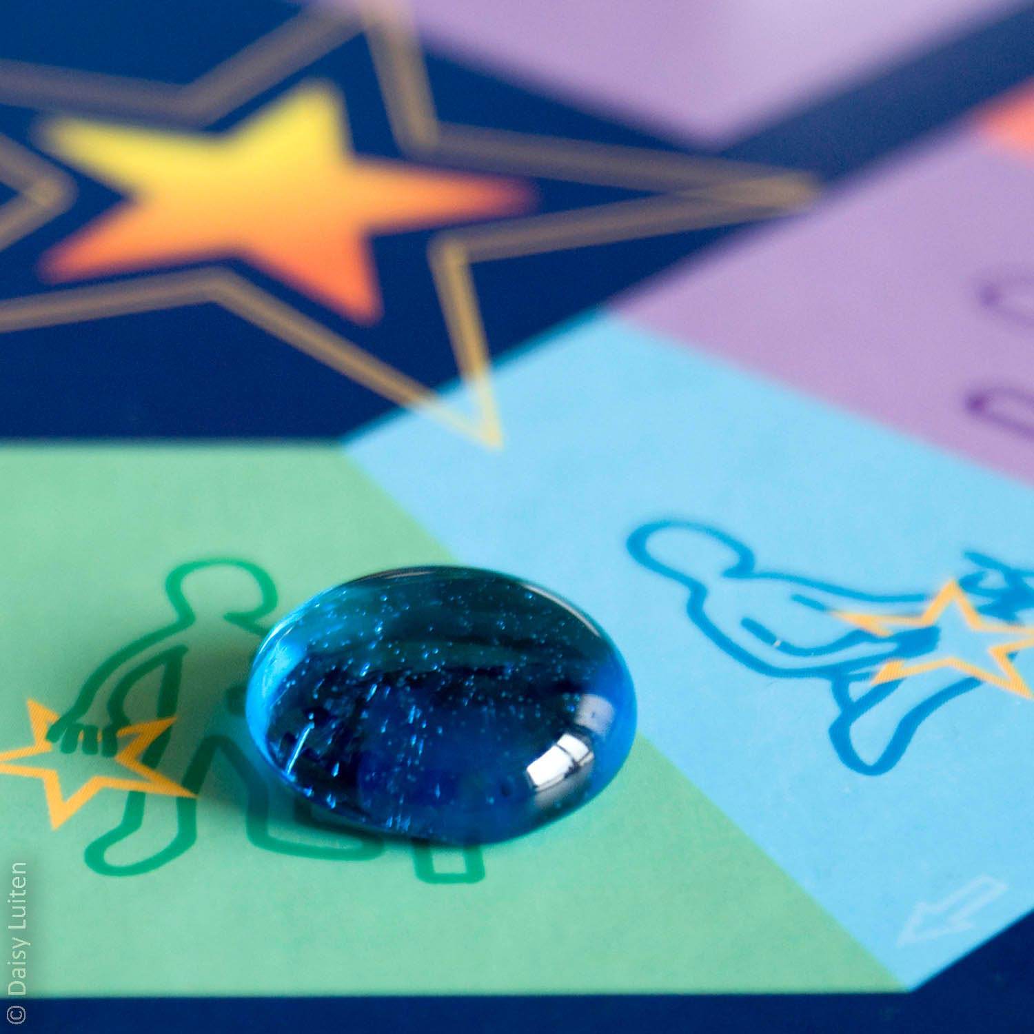 De pion, een glazen blauwe traan ofwel parel, ligt op een groen vakje van het spelbord Alle Sterren Van De Hemel. De gekleurde vakjes met daarin een afbeelding van een poppetje staan symbool voor de categorieën van het therapeutische spel.