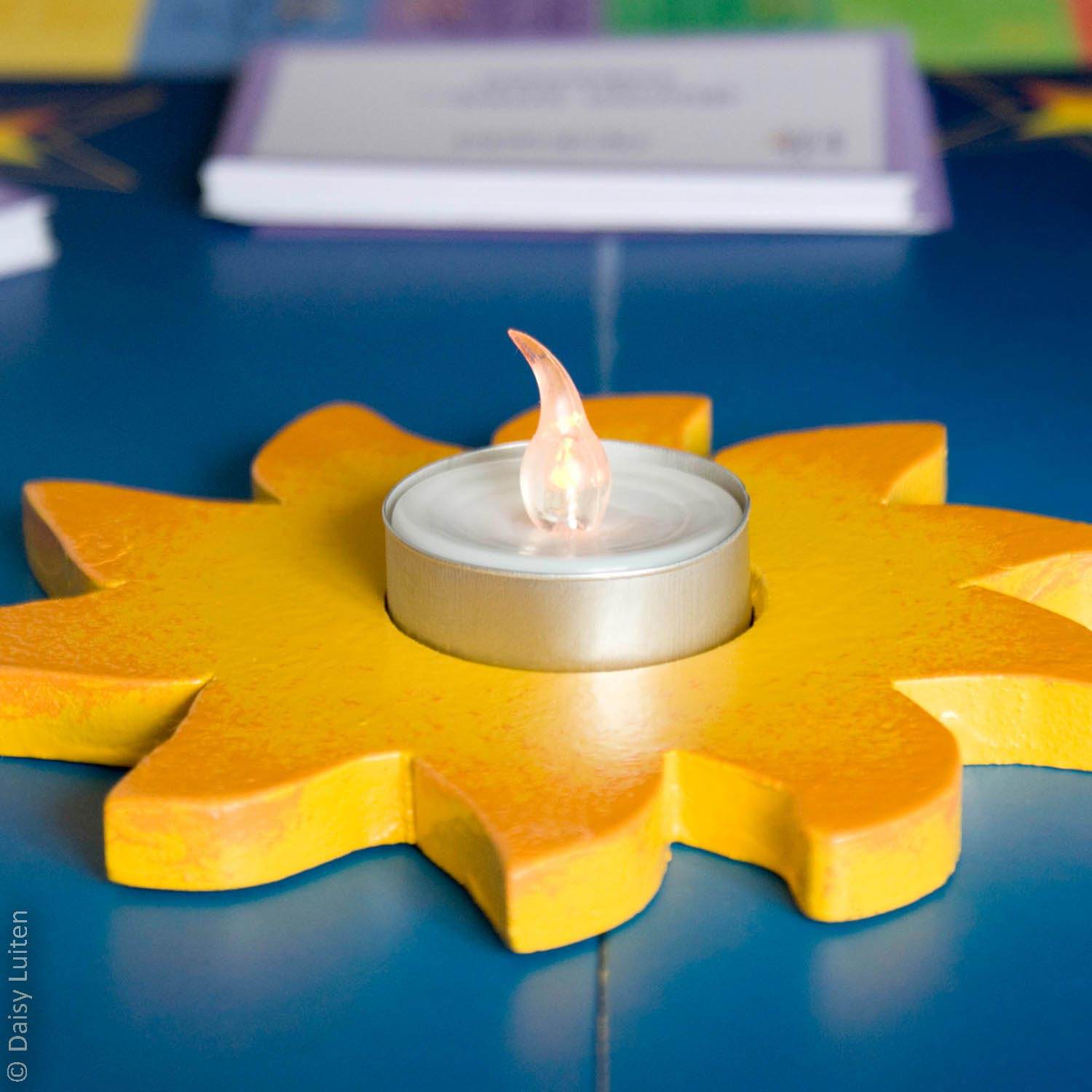 Midden op het blauwe Alle Sterren Van De Hemel spelbord, bevindt zich een gele houten zon met daarin in led-theelichtje. Het lichtje brandt. Op de achtergrond ligt een stapeltje opdrachtkaartjes.