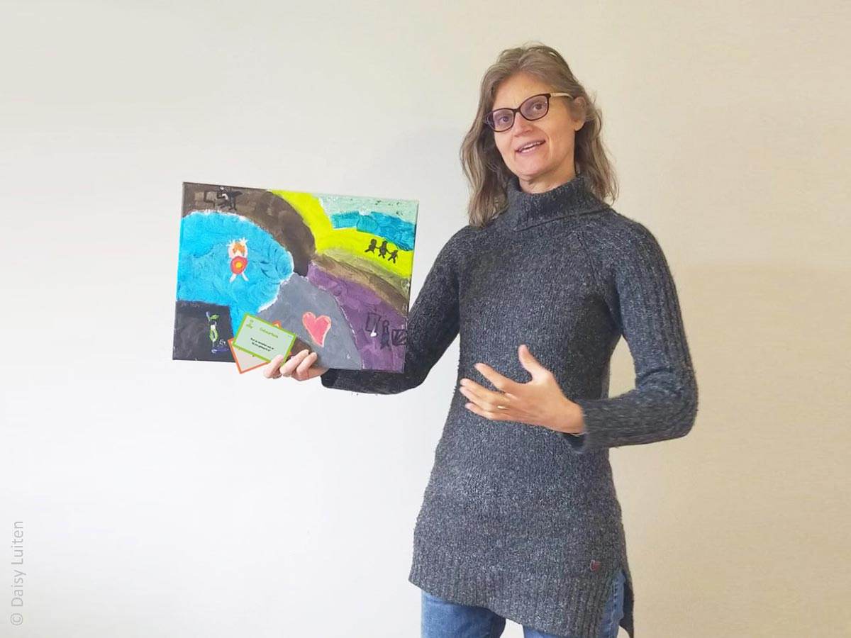 Training_therapeutische_tool_bordspel_Ons-Bestaan-Op-Een-Bordje_creatief_verdiepend_2_Daisy-Luiten1200x900 Daisy Luiten, een vrouw met donkerblond halflang haar en een bril, houdt tijdens het vertellen een schilderij en wat vragenkaartjes in de hand. Daisy geeft hier de creatieve verdiepende training rond haar tool. Een inspirerende training waarbij je in één dag fijne nieuwe werkvormen ontdekt. Om verrassend meer uit Ons Bestaan Op Een Bordje te halen.