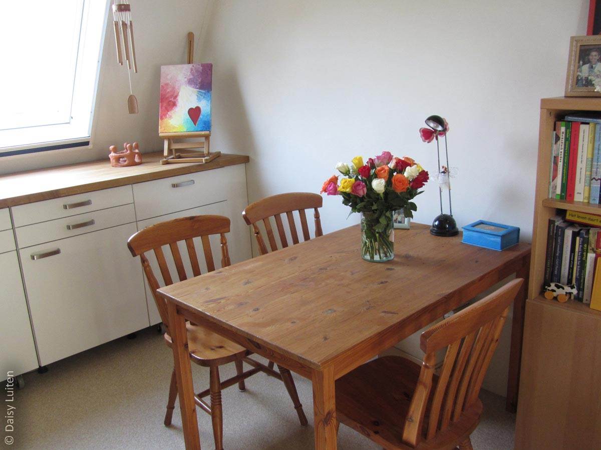 De praktijk van Daisy Luiten oogt warm, door de crème kleurige muren en de houten meubels. Er staat een tafel tegen de muur met 3 stoelen. Een schildering in de achtergrond en een grote bos kleurige rozen op tafel nodigen uit naar binnen te stappen. Rechts van de tafel staat een boekenkast. 