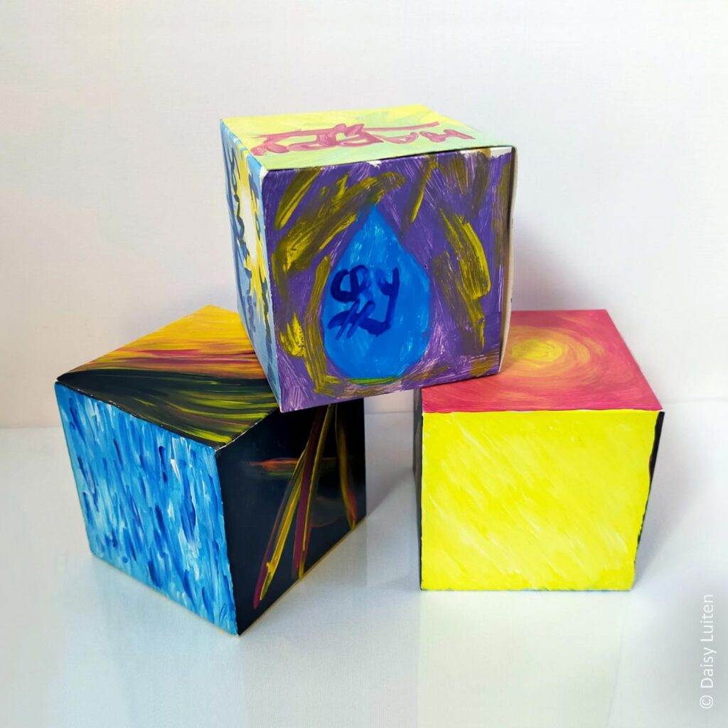 Gevoelens-dobbelstenen. Drie kleurrijk geschilderde kartonnen kubussen. Twee onder en één er bovenop. Op de bovenste staat een blauwe traan geschilderd met 'cry' erin. Op de andere dobbelstenen is onder andere een geel vlak, een blauwvlak en een zwart vlak met geel-rode schrepen te zien.